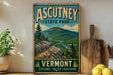 a vintage-style poster of Ascutney State Park, with a mountain landscape and a path leading through the forest. The poster is displayed on a wall, and there are some kitchen utensils, such as a cutting board and a knife, as well as some fruits, such as lemons, in the foreground.