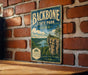 A vintage-style metal sign for Backbone State Park in Iowa, featuring a scenic landscape with a cliff overlooking a lake.