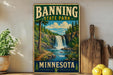 A vintage-style sign for Banning State Park in Minnesota, featuring a scenic illustration of a waterfall surrounded by pine trees and a path leading to the waterfall.