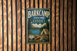 A vintage-style metal sign for Barkcamp State Park in Ohio, featuring a scenic landscape with pine trees, a lake, and a tent, mounted on a wooden wall.