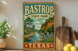 A vintage-style sign for Bastrop State Park in Texas, featuring a scenic illustration of a river, trees, and a cliff.