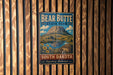 A vintage metal sign for Bear Butte State Park in South Dakota, featuring a mountain landscape with a lake and sunflowers.