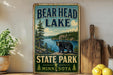 A vintage-style metal sign for Bear Head Lake, featuring a bear and a scenic landscape, is displayed on a shelf next to a wooden cutting board and some lemons.