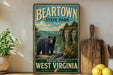 A vintage-style poster of a bear in a mountainous landscape, with the text "BEARTOWN STATE PARK" and "WILD AND WONDERFUL WEST VIRGINIA" printed on it. The poster is displayed on a wall, with a wooden cutting board and a potted plant to the left, and a bunch of lemons to the right.