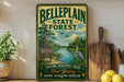 A vintage-style metal sign for Belleplain State Forest, featuring a scenic illustration of a lake, trees, and birds, along with the text "Belleplain State Forest" and "Explore, Discover, Enjoy New Jersey Nature, Recreation, Adventure".