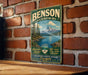 A vintage metal sign for Benson State Recreation Area, featuring a mountain landscape and a river, is displayed on a brick wall.