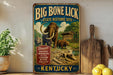 A vintage-style metal sign advertising the Big Bone Lick State Historic Site in Kentucky, featuring an illustration of a prehistoric scene with a large elephant, a bear, and a dinosaur.