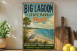 A vintage-style sign for Big Lagoon State Park in Florida, featuring a beach scene with a crane, palm trees, and a sailboat.