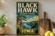 A vintage-style sign for Black Hawk State Park in Iowa, featuring a scenic landscape with a river, cliffs, and a bird in flight.