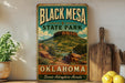 A vintage-style sign for Black Mesa State Park in Oklahoma, featuring a scenic mountain landscape with a river and desert terrain.