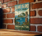 A vintage-style metal sign for " Blake Island State Park" is displayed on a brick wall, featuring a scenic image of the park and a boat on the water.