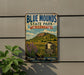 A vintage-style metal sign for Blue Mounds State Park in Minnesota, featuring a scenic landscape with mountains, flowers, and a pine tree.