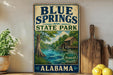 A vintage-style sign for Blue Springs State Park in Alabama, featuring a scenic illustration of a river, trees, and a wooden cabin.