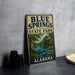 A vintage-style sign for Blue Springs State Park in Alabama, featuring a scenic illustration of a river, trees, and a wooden cabin.