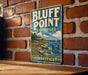 A vintage-style metal sign for Bluff Point State Park in Connecticut, featuring a scenic coastal landscape with a sailboat and rocky cliffs.
