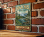 A vintage-style sign for Bogachiel State Park in Washington, featuring a scenic mountain landscape with a river and pine trees.