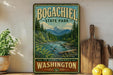 A vintage-style sign for Bogachiel State Park, featuring a scenic mountain landscape with a river and pine trees, is displayed on a wooden shelf next to a wooden cutting board and some lemons.
