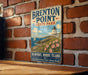 A vintage-style sign for "Brenton Point State Park" hanging on a brick wall, featuring a lighthouse and a coastal landscape with flowers.