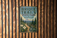 A vintage metal sign for Bridle Trails State Park in Washington, featuring a scenic mountain landscape and a trail leading through the woods.