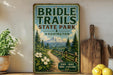 A vintage-style metal sign for Briddle Trails State Park in Washington, featuring a scenic mountain landscape with a trail and flowers.