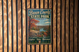 A vintage metal sign for Brown County State Park in Indiana, featuring a scenic landscape with a river and autumn foliage.