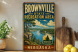 A vintage-style sign for the Brownville State Recreation Area in Nebraska, featuring a scenic landscape with a river, mountains, and a pine tree.