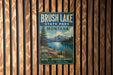 A vintage metal sign for Brush Lake State Park in Montana, featuring a scenic landscape with a lake, mountains, and pine trees.