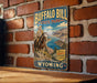 A vintage-style metal sign for Buffalo Bill State Park in Wyoming, featuring a cowboy on horseback and a scenic mountain landscape.