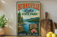 A vintage-style metal sign for Burnsville Lake State Park, featuring a scenic landscape with mountains, a lake, and pine trees. The sign also includes the text "Wild and Wonderful West Virginia" and "Adventure Relax Explore".