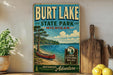 A vintage-style sign for Burt Lake State Park in Michigan, featuring a canoe on a lake and a pine tree, with the text "Burt Lake State Park - Michigan" and "Look Explore Relax" on it. The sign is displayed on a shelf next to a potted plant and a cutting board, with a lemon and a knife visible in the background.