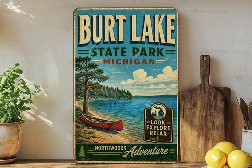 A vintage-style sign for Burt Lake State Park in Michigan, featuring a canoe on a lake and a pine tree, with the text "Burt Lake State Park - Michigan" and "Look Explore Relax" on it. The sign is displayed on a shelf next to a potted plant and a cutting board, with a lemon and a knife visible in the background.