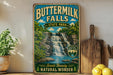A vintage-style sign for Buttermilk Falls State Park, featuring a waterfall and lush greenery, is displayed on a wooden shelf next to a potted plant and a wooden cutting board.