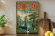 a vintage-style sign for Caddo Lake, Texas, with a painting of a boat on the lake and a tree with hanging roots. The sign is displayed on a shelf next to a wooden cutting board and a potted plant.