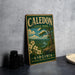 A vintage metal sign for Caledon State Park in Virginia, featuring a scenic landscape with a river and trees, is displayed on a gray countertop next to a stack of black camera lenses.