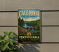 A vintage-style metal sign for Caledonia State Park in Pennsylvania, featuring a scenic landscape with a lake, forest, and waterfall.