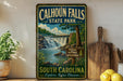 A vintage-style metal sign for Calhoun Falls State Park, featuring a waterfall and lush greenery, is displayed on a shelf next to a wooden cutting board and some lemons.