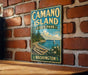 A vintage-style metal sign for Camano Island State Park in Washington, featuring a scenic landscape with a sailboat on the water and a forested shoreline.