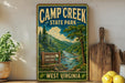 A vintage-style sign for Camp Creek State Park in West Virginia, featuring a scenic illustration of a river and mountains.
