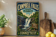 A vintage-style sign for Campbell Falls State Park in Connecticut, featuring a waterfall and lush greenery, is displayed on a wooden shelf next to a potted plant and a cutting board.