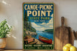 A vintage-style sign for Canoe-Picnic Point State Park in New York, featuring a scenic image of a red canoe on a lake surrounded by pine trees and mountains.