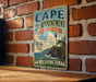 A vintage-style metal sign for Cape Disappointment State Park in Washington, featuring a lighthouse and a coastal landscape.