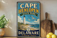 A vintage-style sign for Cape Henlopen State Park in Delaware, featuring a lighthouse and beach scene, is displayed on a shelf next to a wooden cutting board and a potted plant.