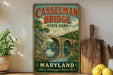 A vintage-style metal sign for Casselman Bridge State Park in Maryland, featuring a scenic illustration of the bridge over a river.