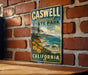 A vintage metal sign for Caswell Memorial State Park in California, featuring a scenic coastal landscape with mountains, trees, and a beach.