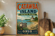 A vintage-style sign for Catawba Island State Park in Ohio, featuring a scenic illustration of a lighthouse, a boat, and a beach.