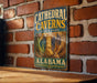 A vintage metal sign for Cathedral Caves State Park in Alabama is displayed on a brick wall.