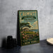 A vintage metal sign for Caumsett State Historic Park Preserve, featuring a scenic illustration of a house and landscape, is displayed on a gray countertop next to a stack of black camera lenses.
