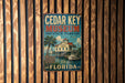 A vintage-style metal sign for the Cedar Key Museum in Florida, featuring a tropical beach scene with palm trees and a boat.