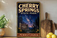 A vintage-style sign for Cherry Springs State Park in Pennsylvania, featuring a night sky scene with stars and a galaxy.