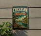 A vintage metal sign for Chickasaw State Park in Alabama, featuring a scenic landscape with a river, trees, and a cabin.